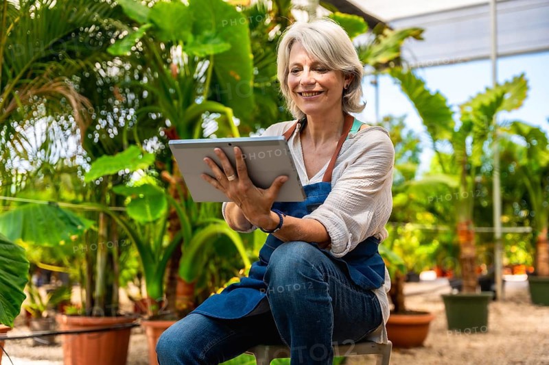 Happy mature woman working in plant nursery and using digital tablet, managing plants and flowers