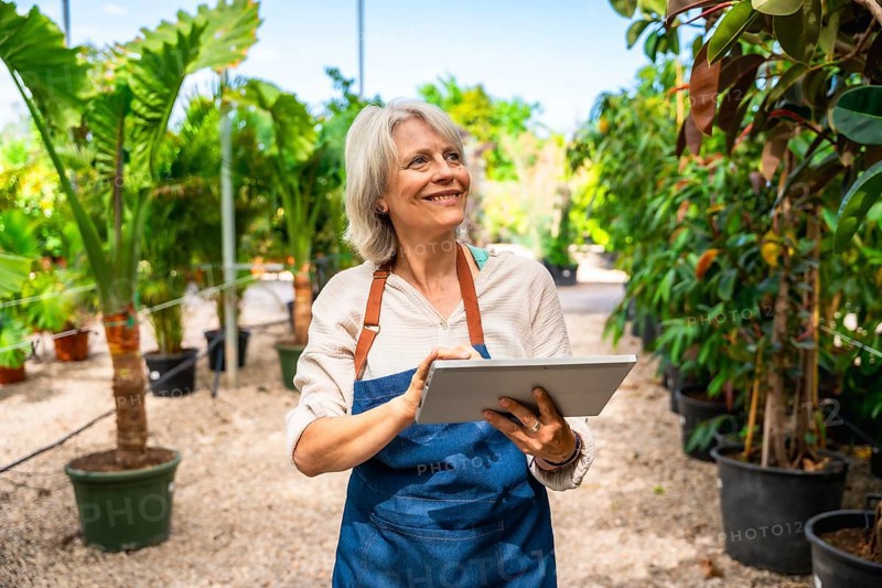 Smiling gardener working in a greenhouse, using a digital tablet while surrounded by vibrant plants and enjoying the sunny day