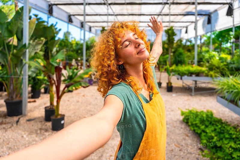 Young woman gardener with curly red hair enjoying the scent of plants, taking a selfie while stretching arms in a greenhouse
