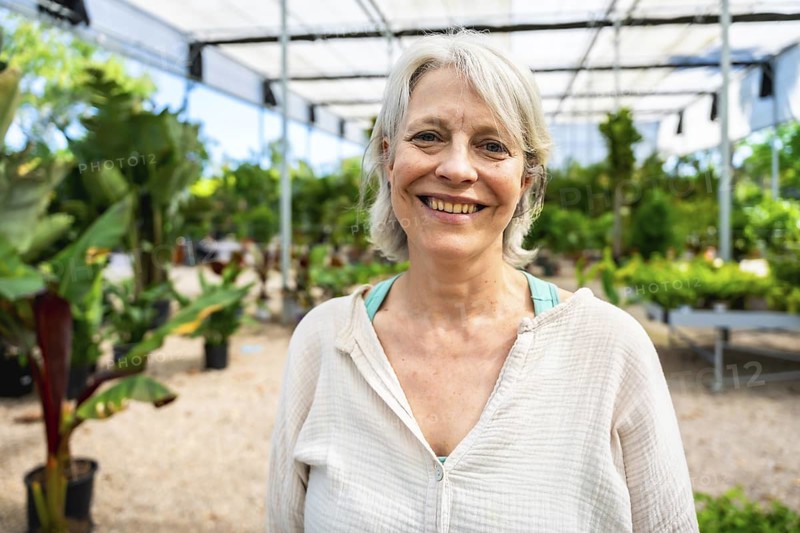 Portrait of a cheerful mature woman working joyfully in a greenhouse, surrounded by vibrant, lush plants and thriving greenery