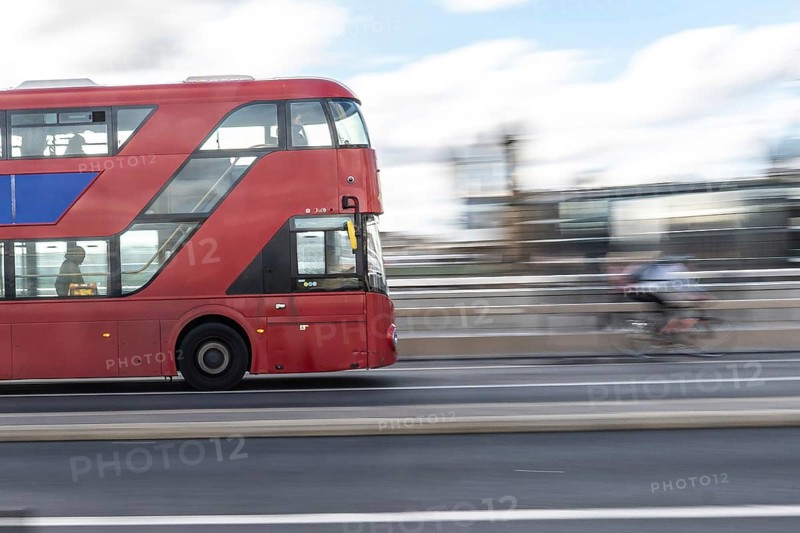 Red double decker bus driving on a london road with a cyclist passing by in the background, capturing the city's dynamic urban life