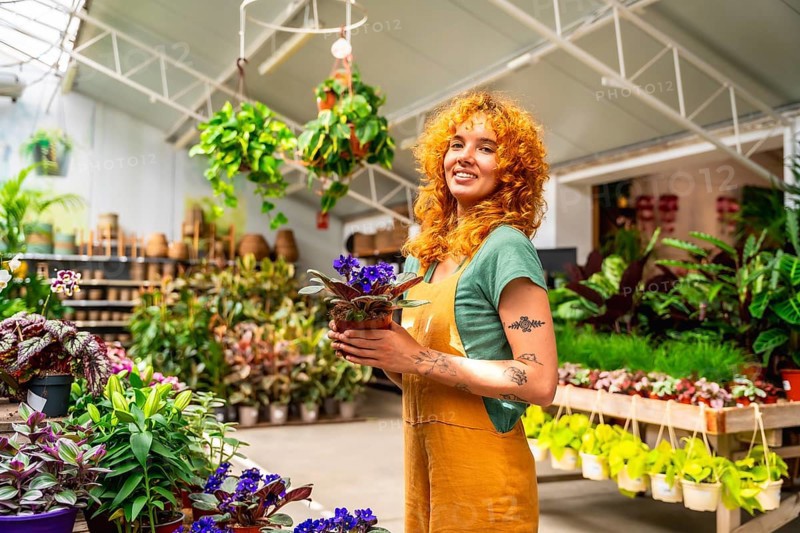 Young saleswoman holding a vibrant potted plant and smiling brightly in a lush greenhouse filled with an array of flourishing plants