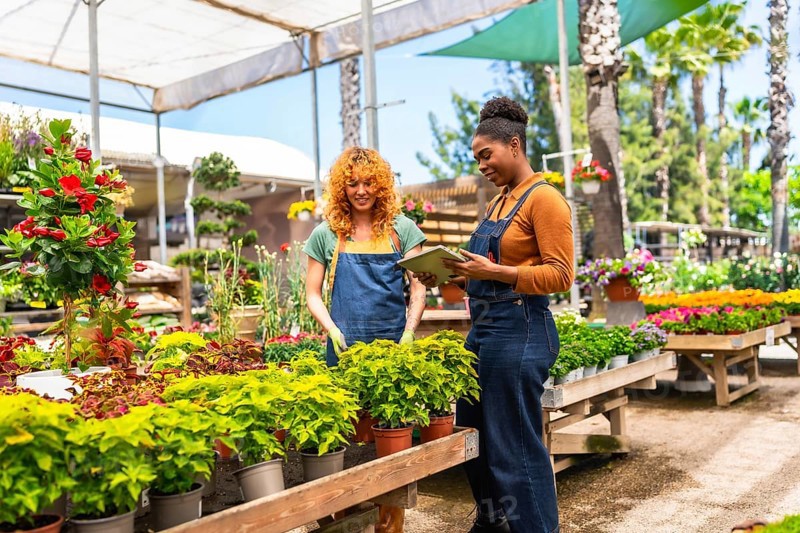 Two women collaborating in a garden center, consulting a digital tablet while discussing various plants and their care