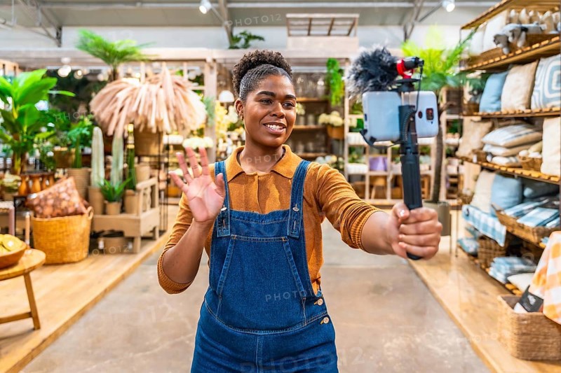 Young saleswoman waving and streaming live video using a smartphone and microphone inside a furniture store