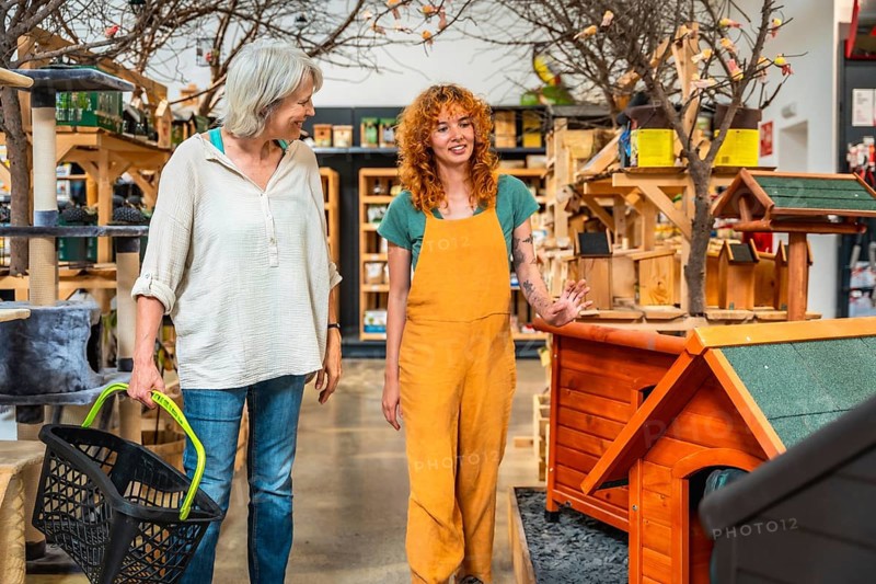 Smiling saleswoman showing wooden dog houses to customer carrying shopping basket in a pet shop