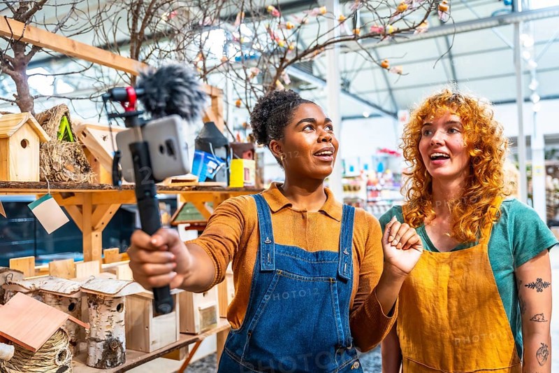 Two young women working in a garden center filming a video for social media using a smartphone with external microphone