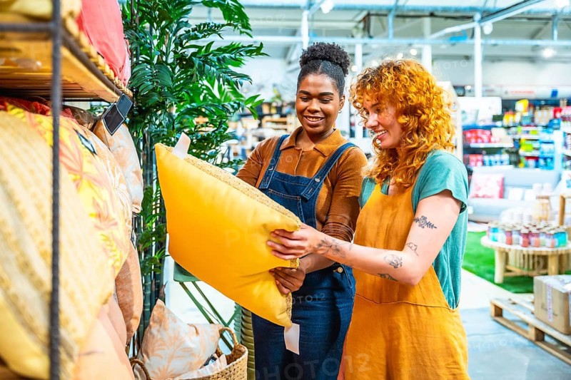 Two cheerful sales assistants collaborating in a home decor store, holding a vibrant yellow pillow while providing excellent customer service