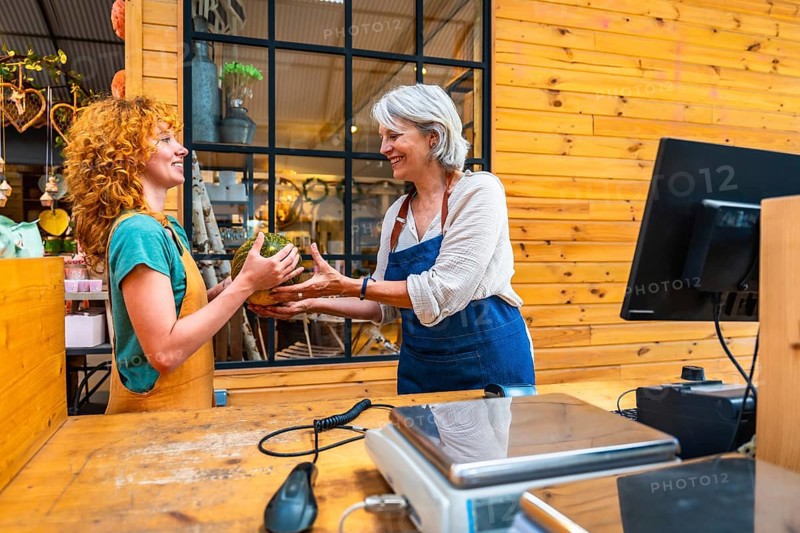 Two women joyfully exchanging a pumpkin in a vibrant flower shop, with a scale and computer prominently displayed on the counter