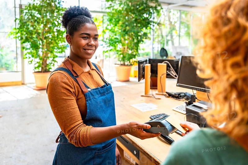Smiling cashier holding pos terminal while customer paying with smartphone using nfc technology in a plant store