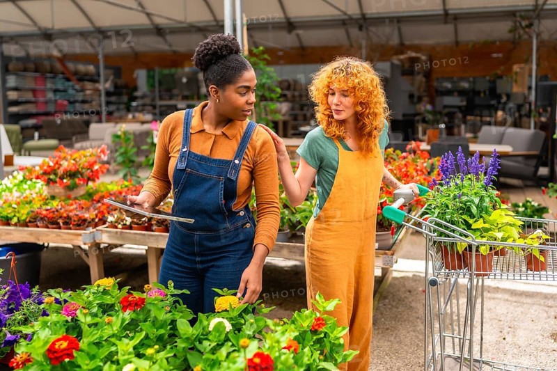 Two women working in a garden center are discussing flowers while using a digital tablet and pushing a shopping cart