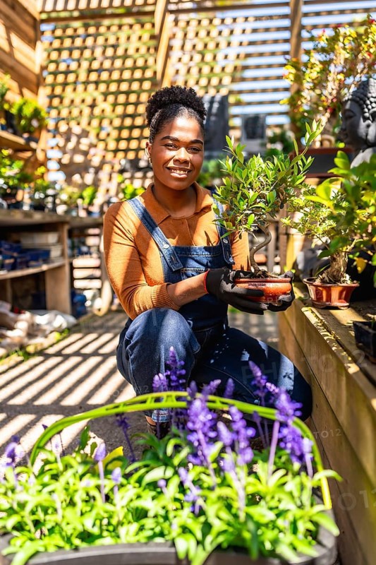 Young woman working in a vibrant greenhouse, holding a small bonsai tree, surrounded by flourishing plants and colorful flowers