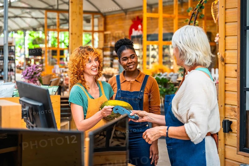 Customer paying with contactless payment in a grocery store, assisted by two smiling shop assistants