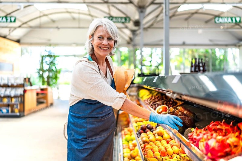 Smiling greengrocer wearing protective gloves, carefully arranging a vibrant display of fresh fruit in a bustling supermarket environment