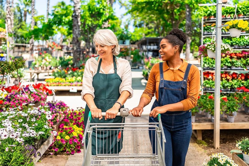 Two female garden center workers pushing an empty shopping cart between rows of colorful potted flowers