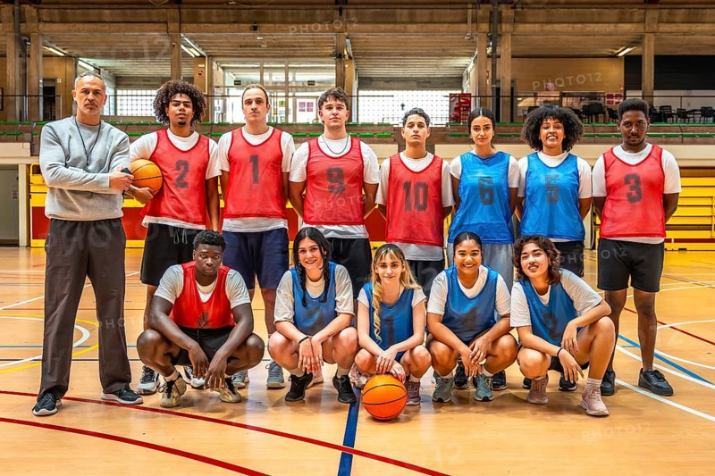 Diverse high school basketball team posing confidently for a group photo with their coach on the court, showcasing teamwork and unity