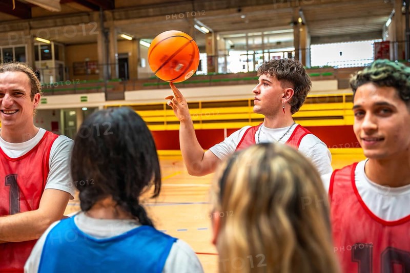 Young basketball player spinning ball on finger, surrounded by teammates in gym, showcasing skill and coordination