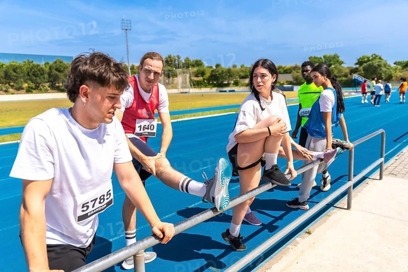 Runners stretching their legs on a railing, preparing for competition on an athletics track under a sunny blue sky