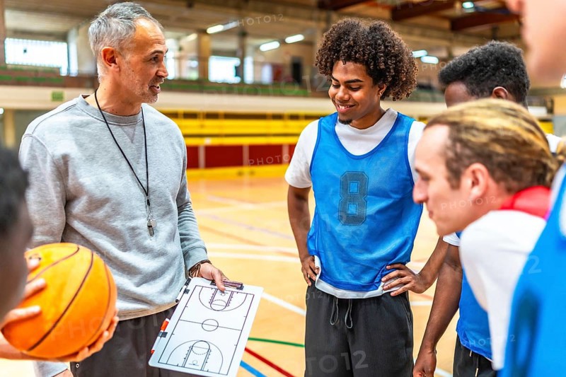 Basketball coach holding ball and clipboard explaining game strategy to team players in gymnasium