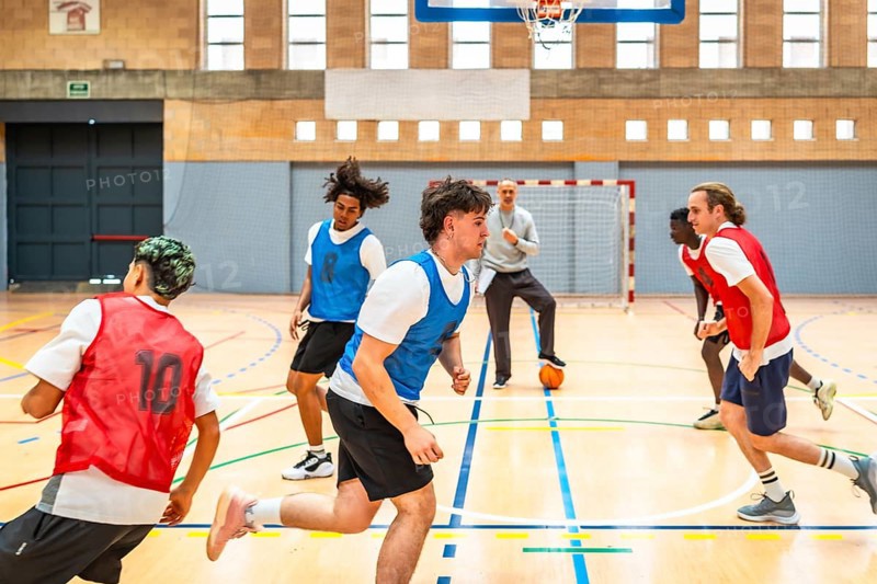 Group of young basketball players running on court during training session with coach watching and ball on floor
