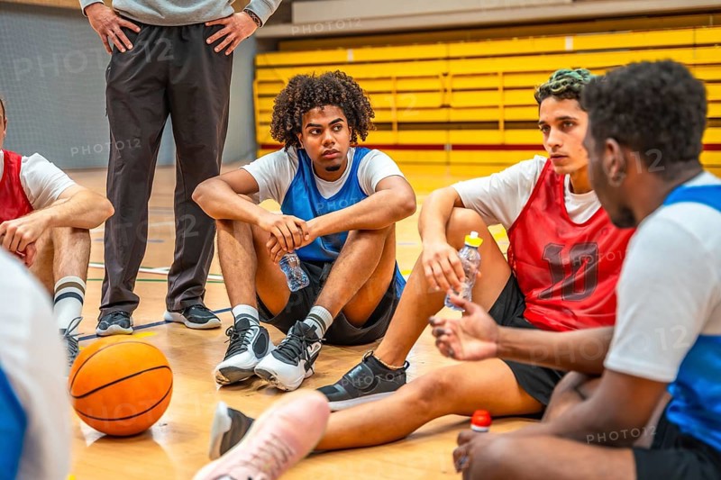 Basketball team resting and drinking water while listening to coach during time out in high school or college gym