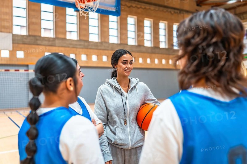 Female basketball coach holding a ball while engaging with her team during an intense training session in the gym, fostering teamwork and strategy