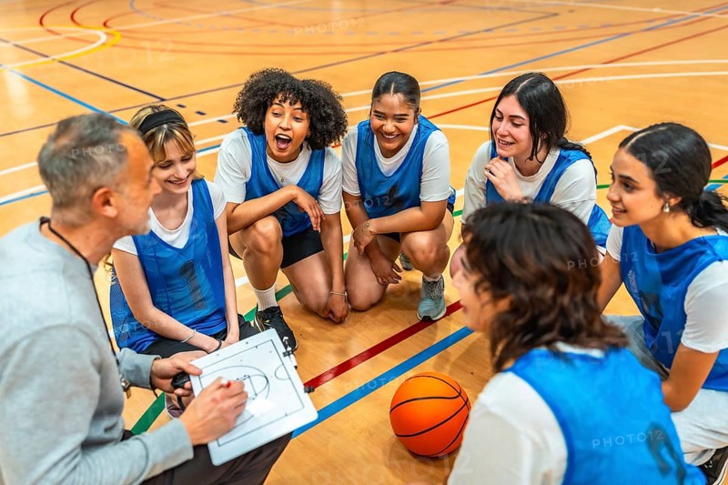 Female basketball team listening to their coach explaining game strategy on a clipboard during a time out