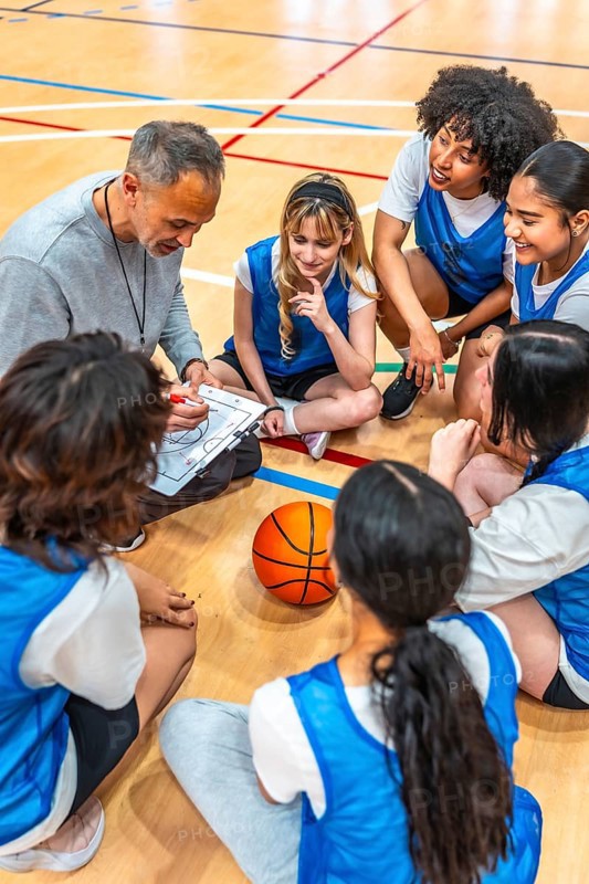 High angle view of a basketball coach drawing a play on a clipboard while sitting on the court with his female high school team