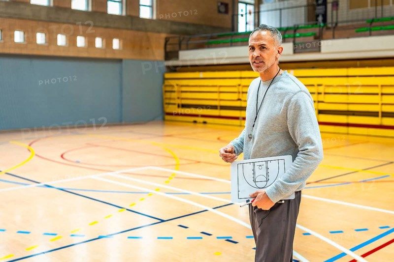 Mature basketball coach holding clipboard with game strategy explaining tactics to team players in indoor court