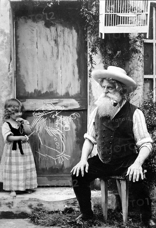 Granddaughter paints her smoking grandpa, 1910, An elderly man with a beard sits in front of a door with a painted chalk drawing, a little girl stands next to him, Historical photo