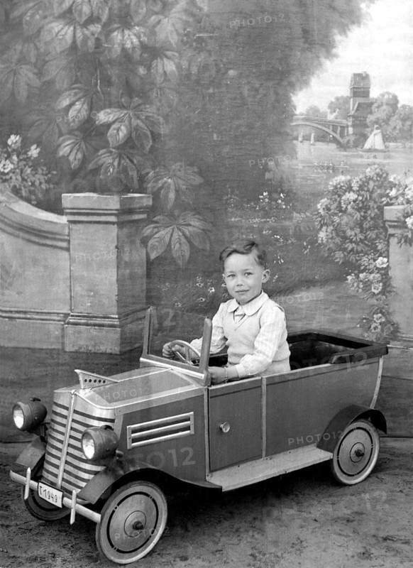 Little boy in car, 1930s, A boy sits in a toy car in front of a picturesque background, Historical photo