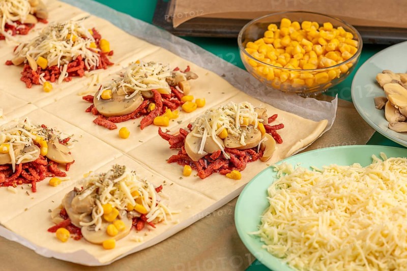 Sliced dough squares with tomato paste, bacon, mushrooms, sweetcorn, cheese and oregano alongside other ingredients for pizza, balls