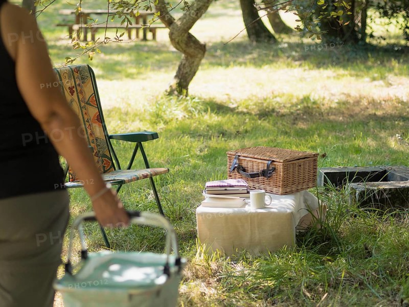 Woman bringing picnic basket to an outdoor setup with a wicker basket and camping chair
