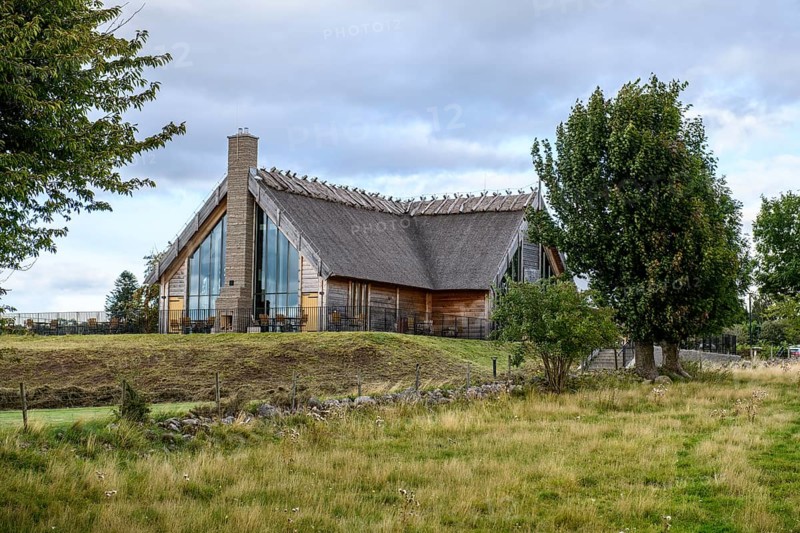 Naturum and Café Doppingen, Naturum Hornborgasjön modern thatched building with glass front and reed roof, which stands in Lake Hornborgasjön and can be reached via footbridges, surrounded by plants under a dramatic sky, Broddetorp, Västra Götalands län, Sweden