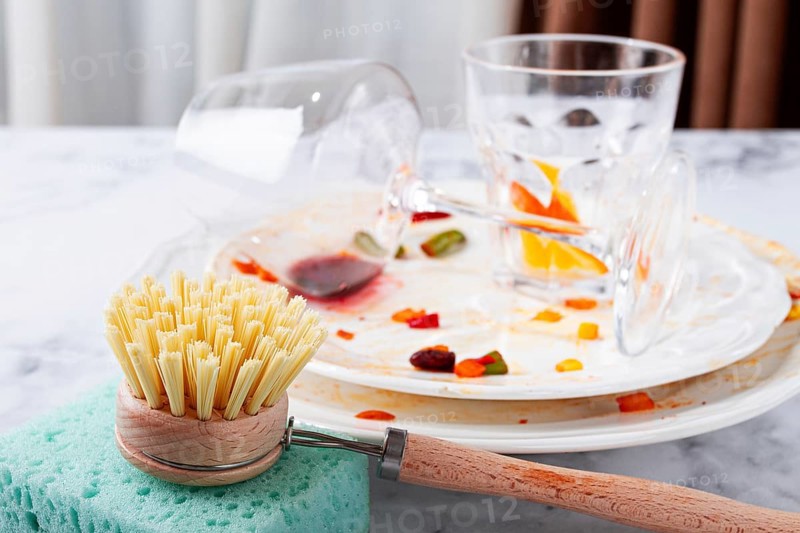 Dishes are stacked on a kitchen counter with food remnants after a meal. A cleaning brush and sponge are ready for washing the plates and glasses