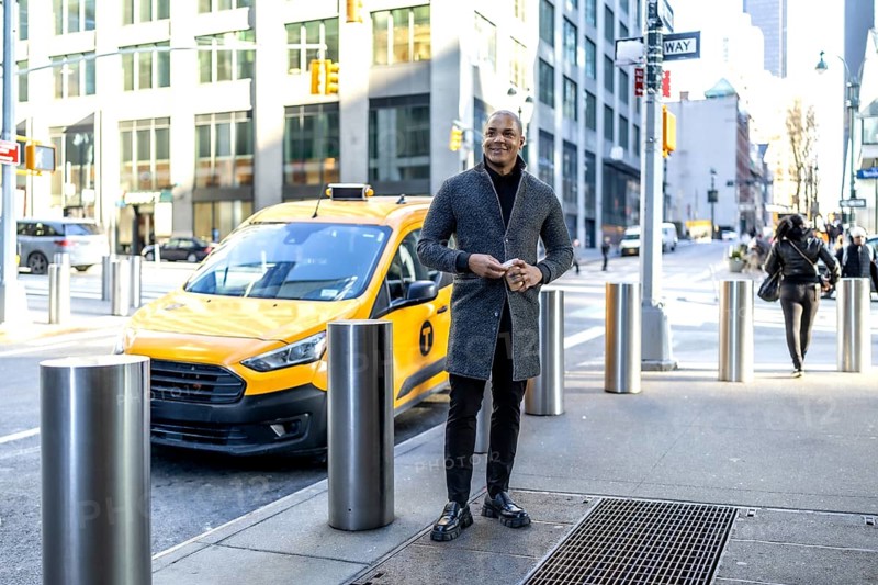 Young businessman stands on a manhattan sidewalk holding a smartphone beside a yellow taxi, smart casual coat signaling confidence during a busy new york commute and urban workday