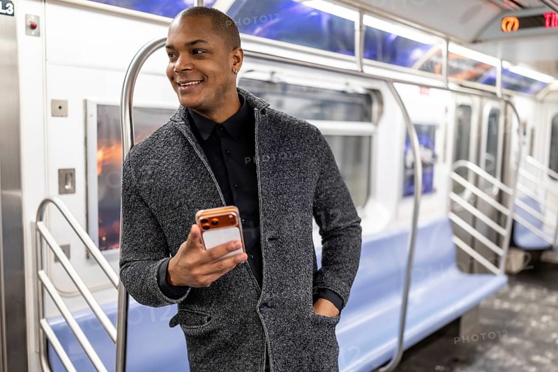 Young african american businessman standing on a subway train in new york city, smiling happily while holding and looking at his mobile phone during an urban commute