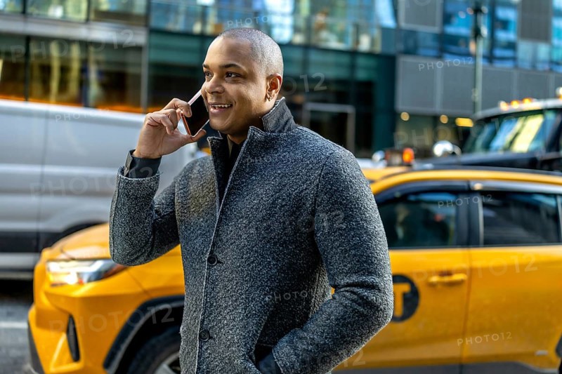 Busy professional man in a winter coat smiling and talking on his smartphone while walking through manhattan with a yellow taxi passing in the busy urban streetscape