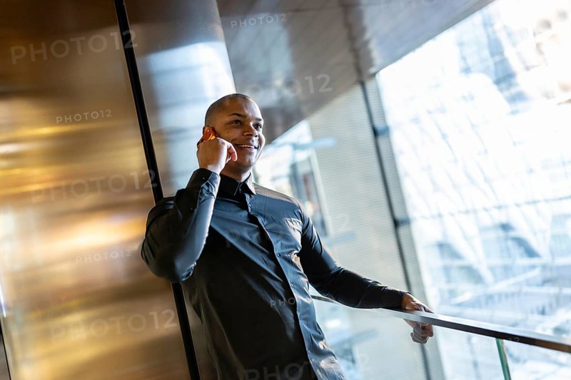 Smiling young professional man is confidently speaking on his cell phone while leaning on a railing inside a sleek urban building, possibly an office or transit hub, conducting business in motion