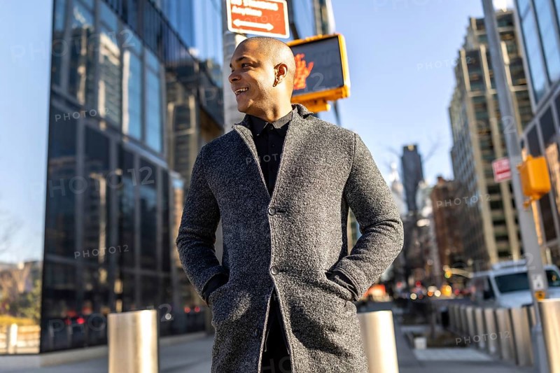 Young man smiling confidently as he walks through manhattan's financial district in smart casual attire, sunny blue sky and modern skyscrapers framing his commute and success