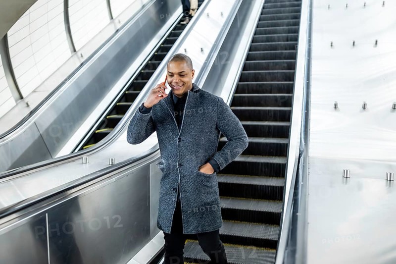 Young man standing on an escalator, holding a cellphone to his ear while smiling, representing urban mobility, communication, and connectivity in a busy metropolitan environment like new york city