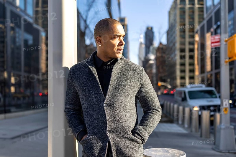 Professional man thinking while standing on a busy new york city street, wearing a stylish gray coat, with modern skyscrapers defining the urban background