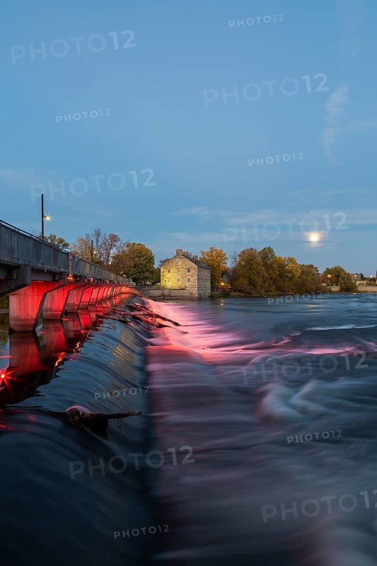Illuminated Moulin Neuf water flow control dam and walkway over Des Mille-Iles river plus New Mill on Ile des Moulins in autumn at dusk, Old Terrebonne, Quebec, Canada