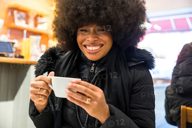 Young black woman joyfully holding a cup of coffee while smiling widely in a cozy cafe, portraying happiness and a cheerful winter moment in new york city