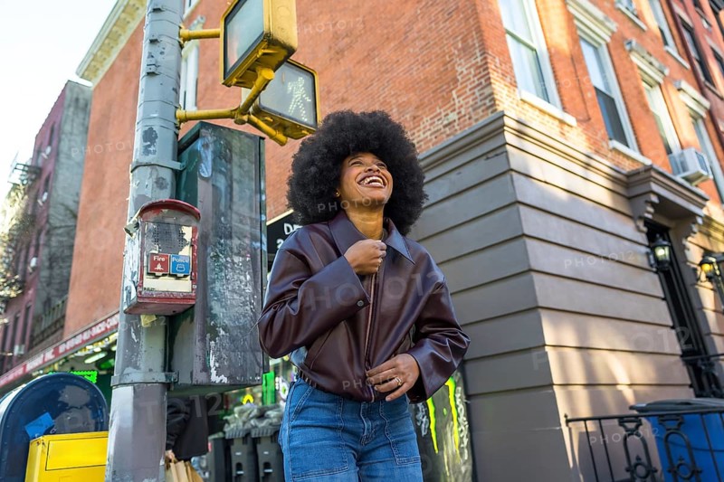 Young african american woman with afro hair smiling and looking up, walking in a vibrant new york city neighborhood street with a brick building and urban infrastructure