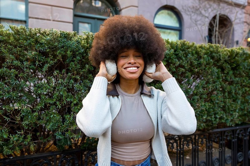 Smiling black woman with large afro hair wearing earmuffs and a sweater, standing outdoors on a winter day in new york city with a green hedge behind her
