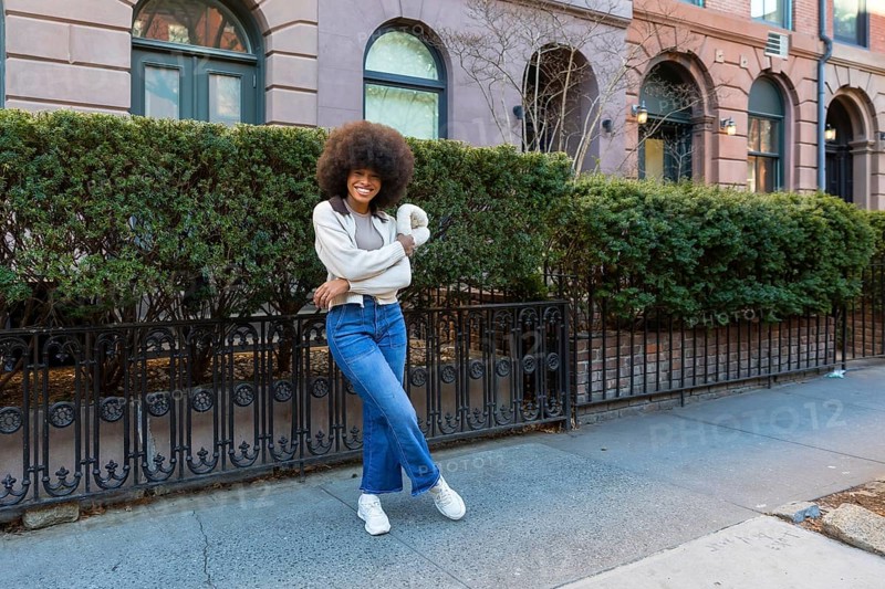 Young black woman with an afro hairstyle smiling confidently while standing on a sidewalk in front of residential buildings, embodying urban lifestyle, joy, and diversity in a city environment