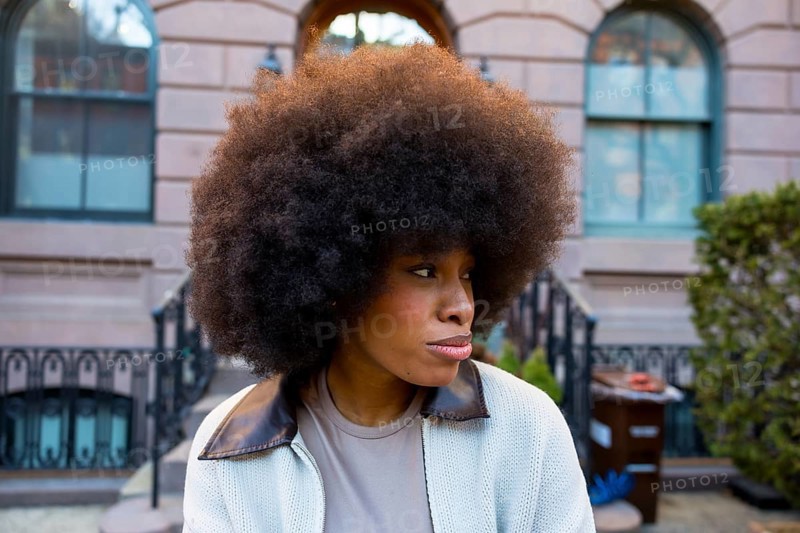 African american woman with a large afro hairstyle looking away, wearing a casual jacket, standing outdoors in front of a manhattan brownstone building representing urban life
