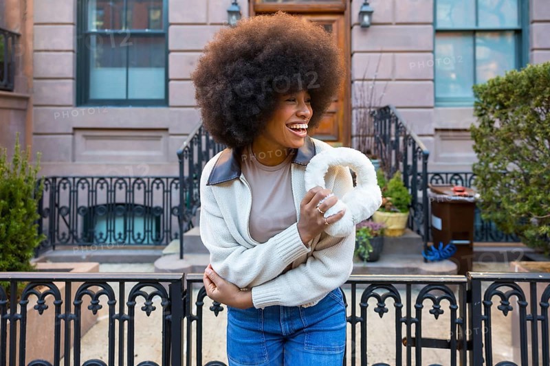 Happy black woman with afro hair laughing, holding furry earmuffs, and wearing jeans with a sweater in an urban setting during cold weather, representing joy, style, and authenticity