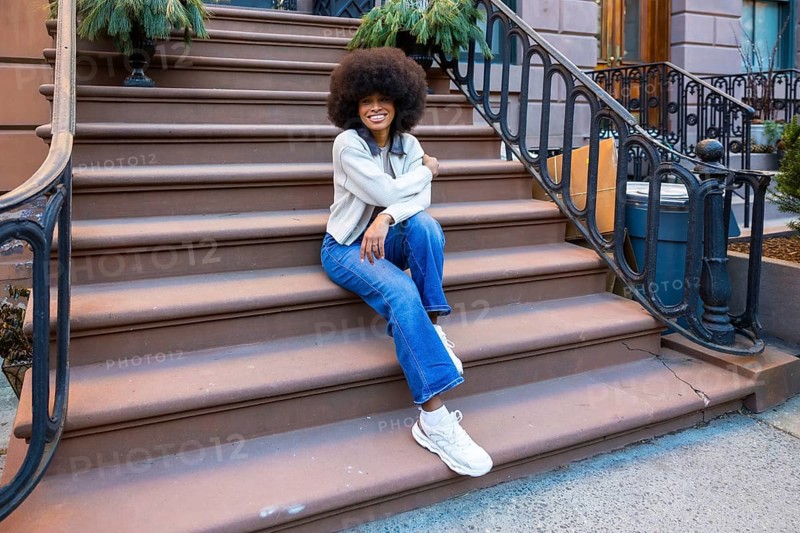 Young african american woman with afro hair sitting on a brownstone stoop in a new york city neighborhood, enjoying urban lifestyle and modern casual fashion