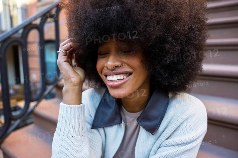 Young black woman laughing with eyes closed and hand in hair, sitting on a brownstone stoop, showcasing joy and spontaneity in an urban new york setting